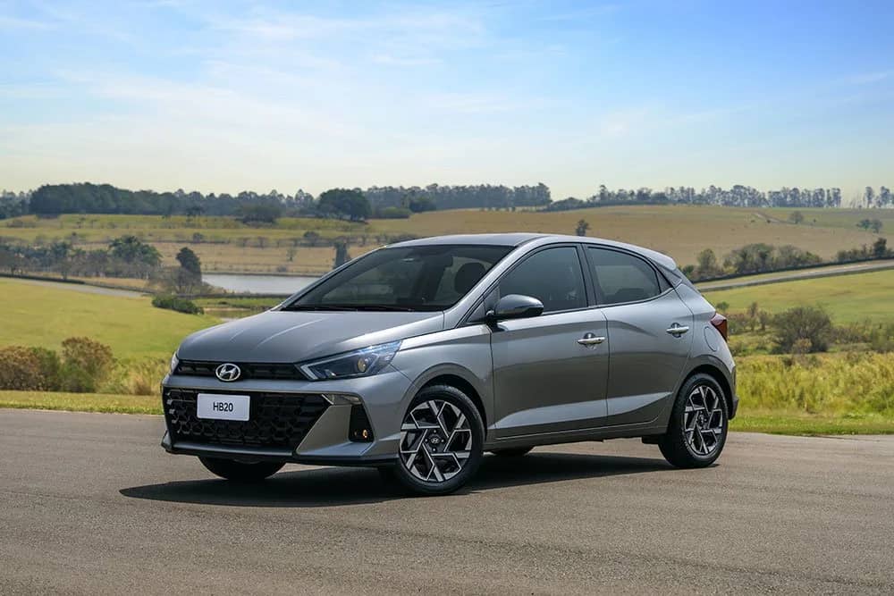 Hyundai HB20 hatch 2027 prata, visto de frente e de lado em uma pista com gramado e árvores ao fundo.