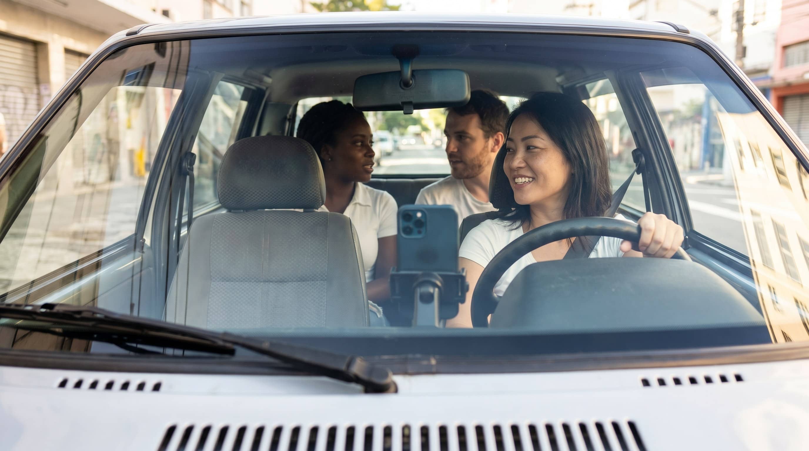 Motorista asiático-brasileira ao volante com celular no console e dois passageiros conversando ao fundo em rua urbana