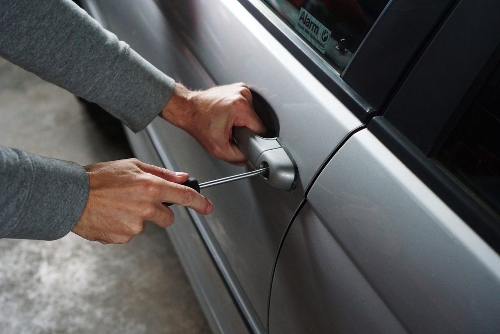 Na imagem há uma pessoa caucasiana, com uma chave de fenda tentando abrir a fechadura de um carro que tem seguro automotivo.