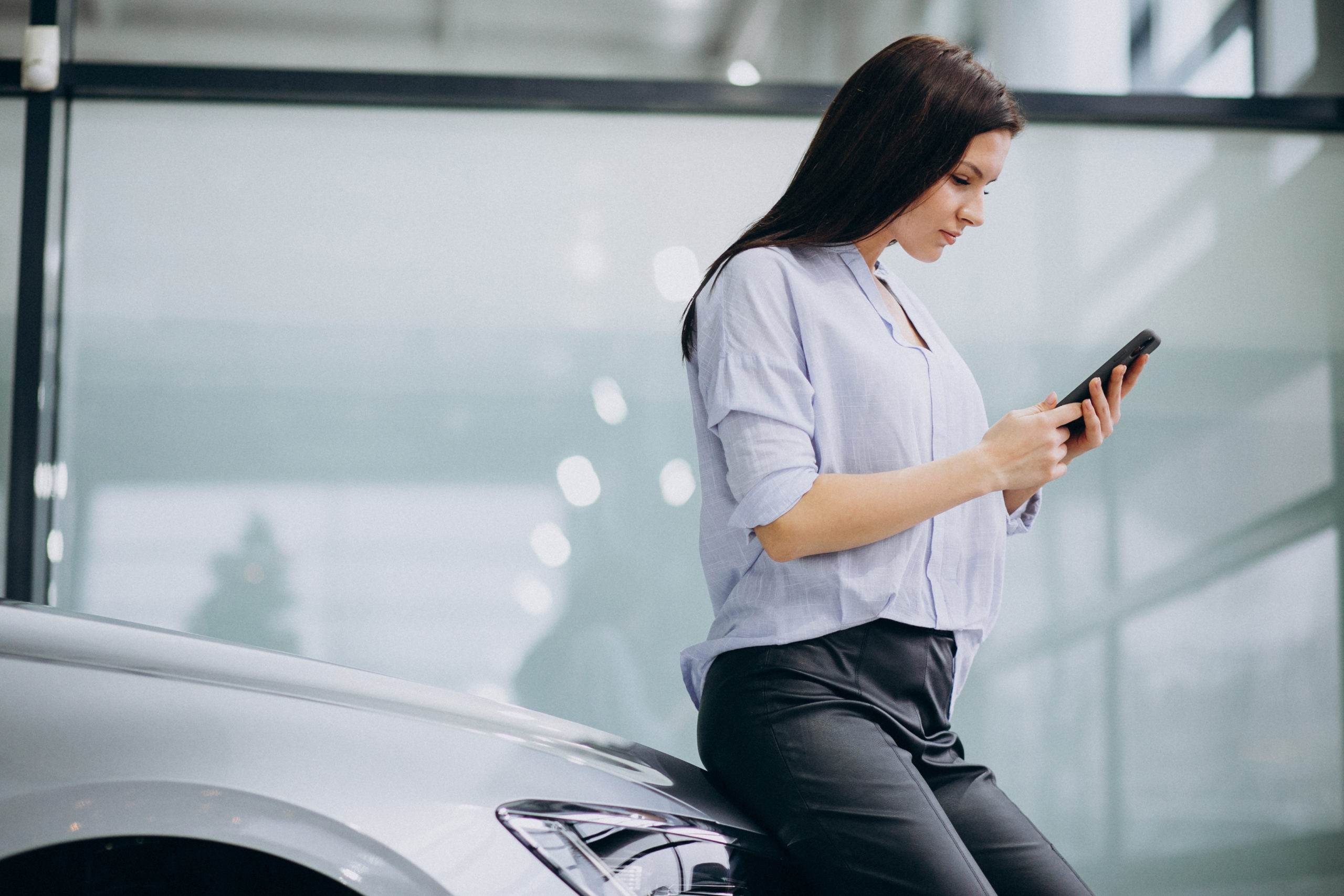 Uma mulher encostada na frente de um carro mexendo em um celular entrando em contato com um app de motorista de aplicativo