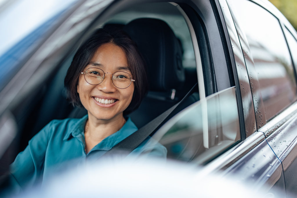 Mulher asiática, com cabelo curto e óculos, blusa azul, dentro de um carro e sorrindo.