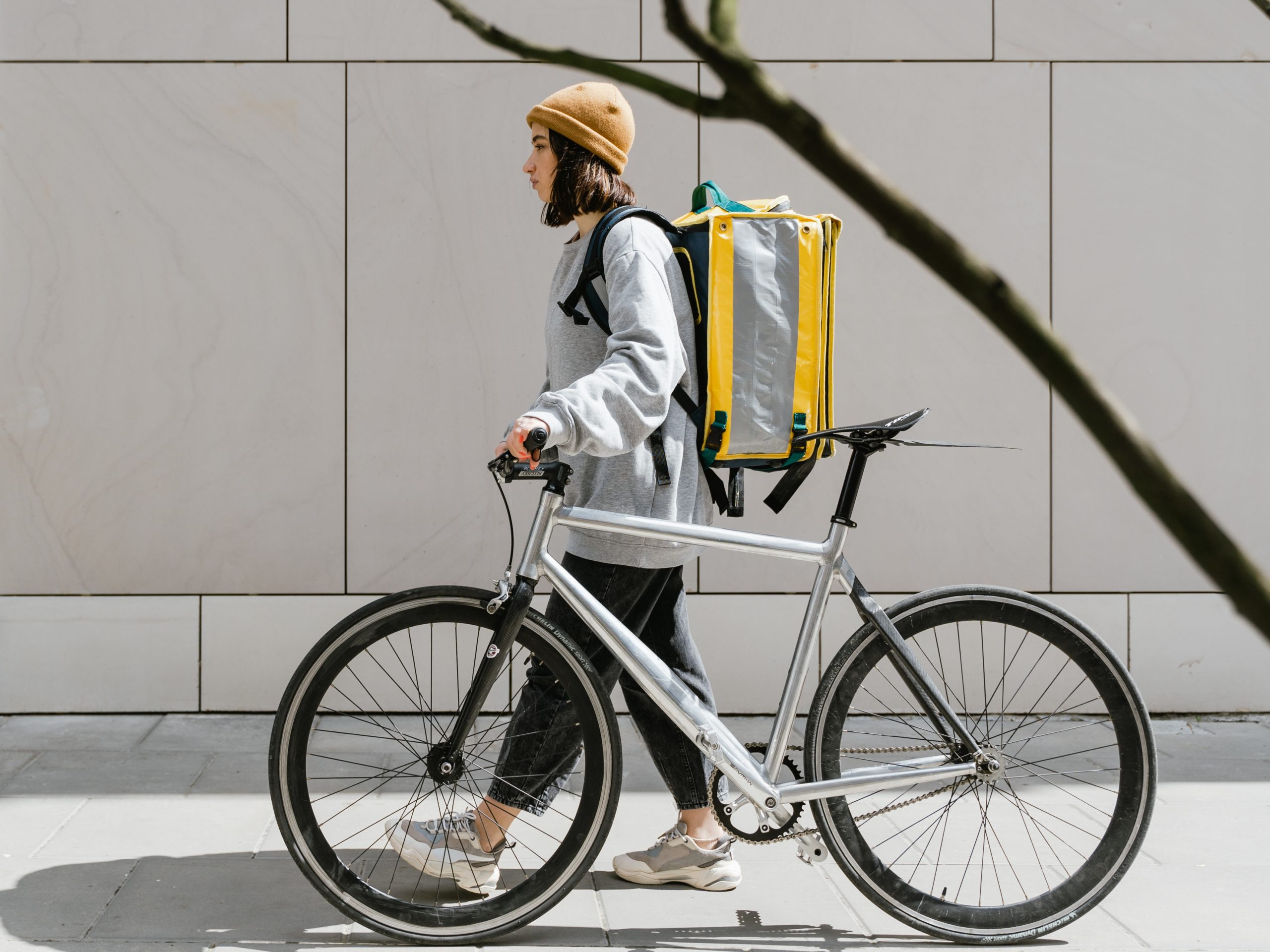 Pessoa andando enquanto leva uma bicicleta e carrega uma mochila amarela de entrega nas costas.