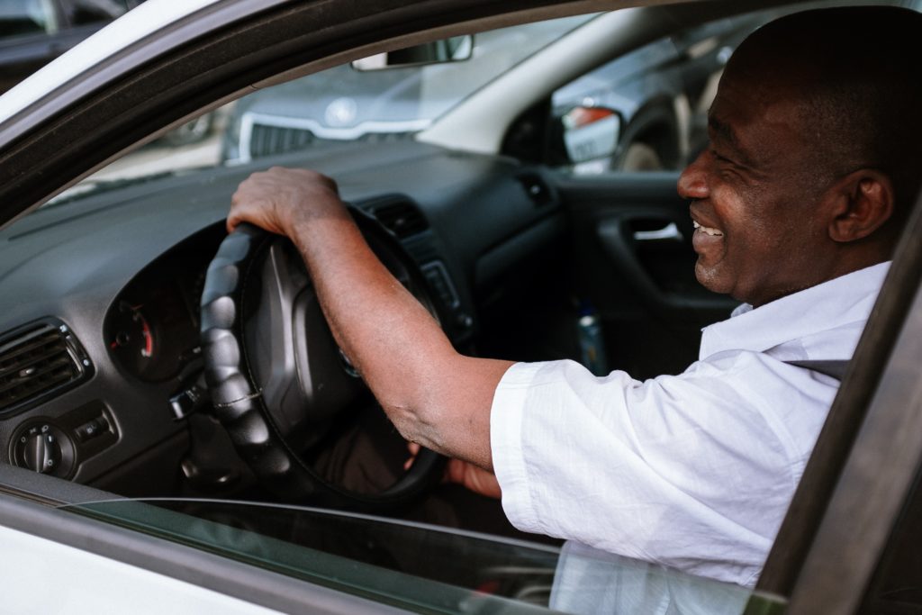 Na imagem há um motorista negro sorrindo, enquanto olha pela janela. Simpatia é um fator chave para ter um serviço de motorista 5 estrelas