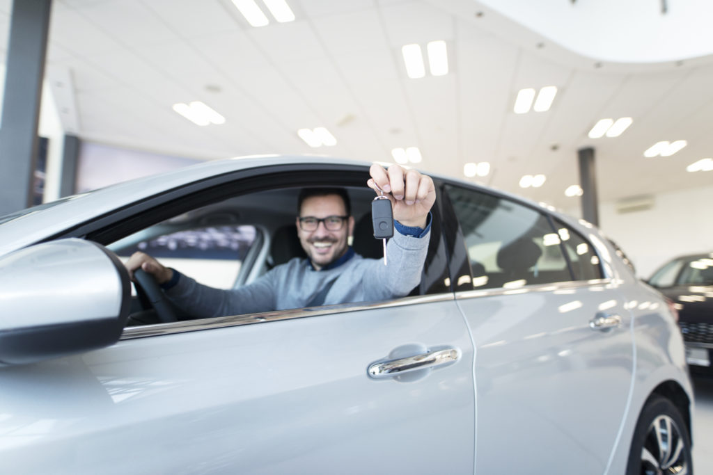Homem dentro de carro, no banco do motorista, segurando a chave do veículo com a mão esquerda para fora do carro enquanto sorri.