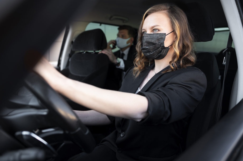 Motorista dirigindo usando máscara. Passageiro no banco de trás, também usando máscara.