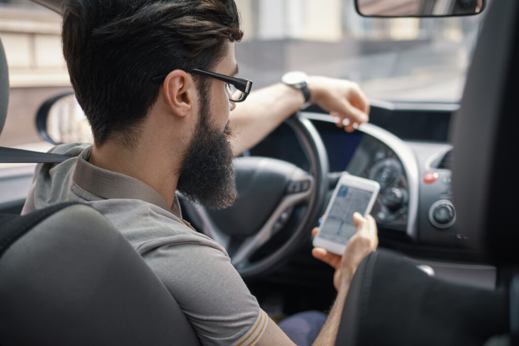 Homem com barba e cabelos escuros vestindo uma camiseta cinza. Ele está dentro do carro verificando a localização no GPS do celular.
