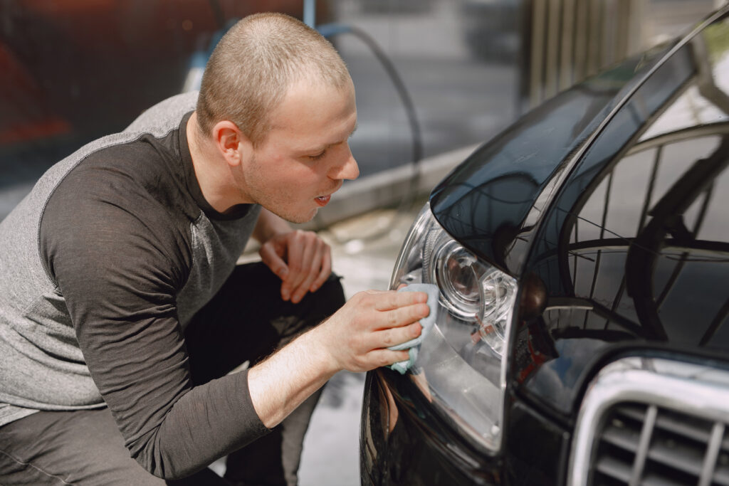 Homem abaixado utilizando uma flanela para limpar o farol dianteiro do carro.
