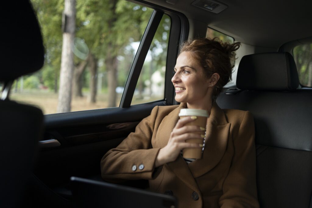 Passageira vestindo casaco marrom está segurando um copo de café enquanto olha a paisagem pela janela do carro.