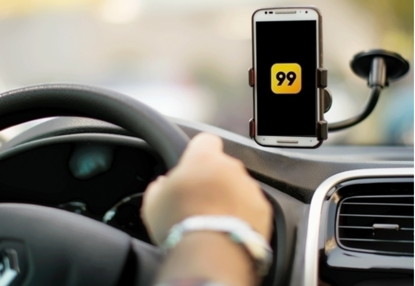Mão em um volante, e, ao lado direito, um celular com a logo da 99, apoiado em um suporte preso ao vidro do carro.