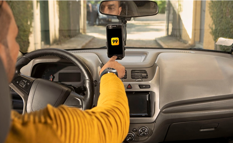 Homem de costas com cabelo curto e blusa amarela, sentado no banco do motorista, com a mão em um celular, e nele, o logo da 99.