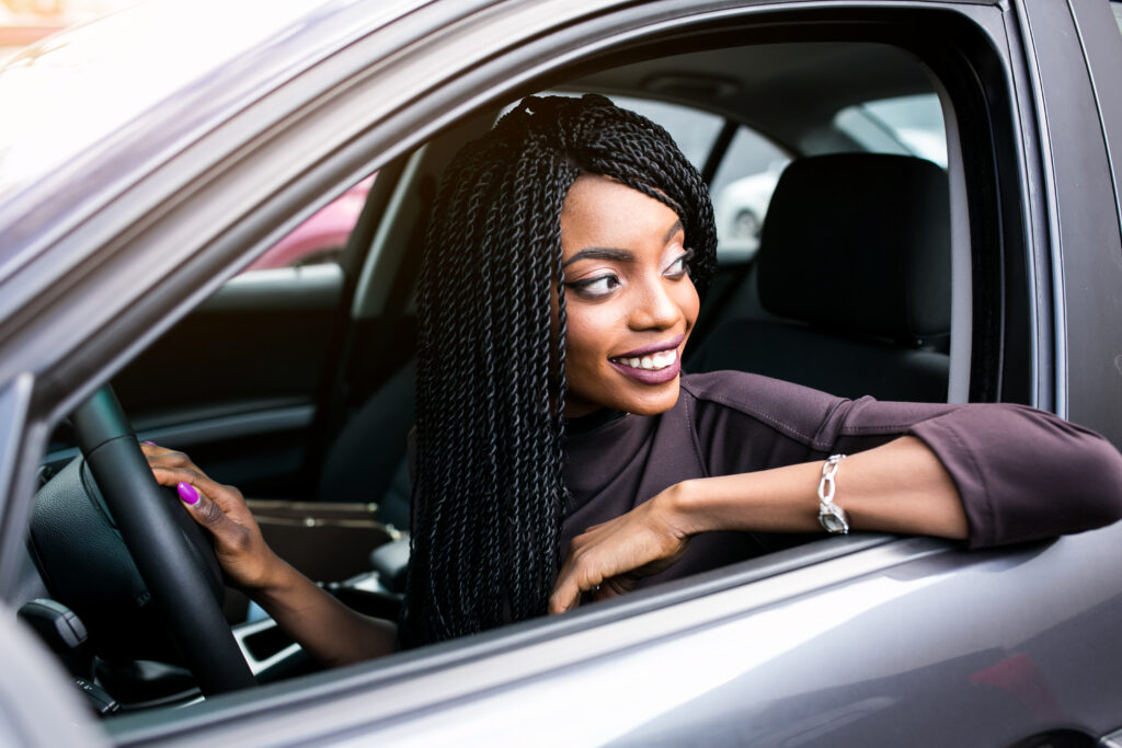 Mulher de cabelo longo preto e camiseta de maga longa marrom, sentada no banco do motorista com uma mão no volante e o outro braço apoiado na janela, olhando para fora do carro pela janela.