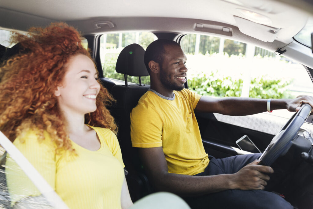 Um homem com cabelo raspado e camiseta amarela sentado no banco do motorista com as mãos no volante. Ao seu lado, no banco do passageiro, uma mulher com cabelo longo e camiseta amarela.