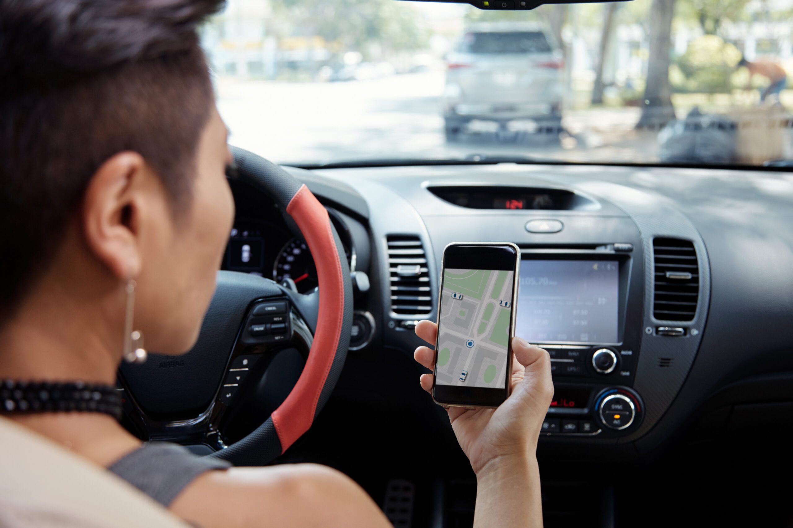 Mulher de costas com cabelo curto, sentada no banco do motorista, olhando para um celular em sua mão em que um mapa está aberto.