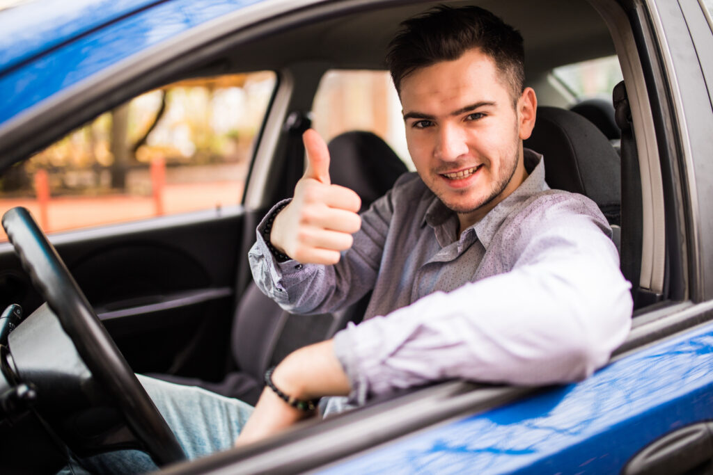 Homem de cabelo curto e camisa cinza sentado no banco do motorista de um carro azul, com um dos braços apoiado na janela e o outro com o polegar levantado, sorrindo.