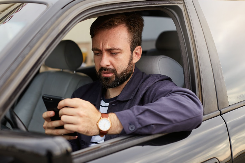 Homem de cabelo curto e barba, vestindo uma camisa preta, sentado no banco do motorista segurando um celular e olhando para ele.