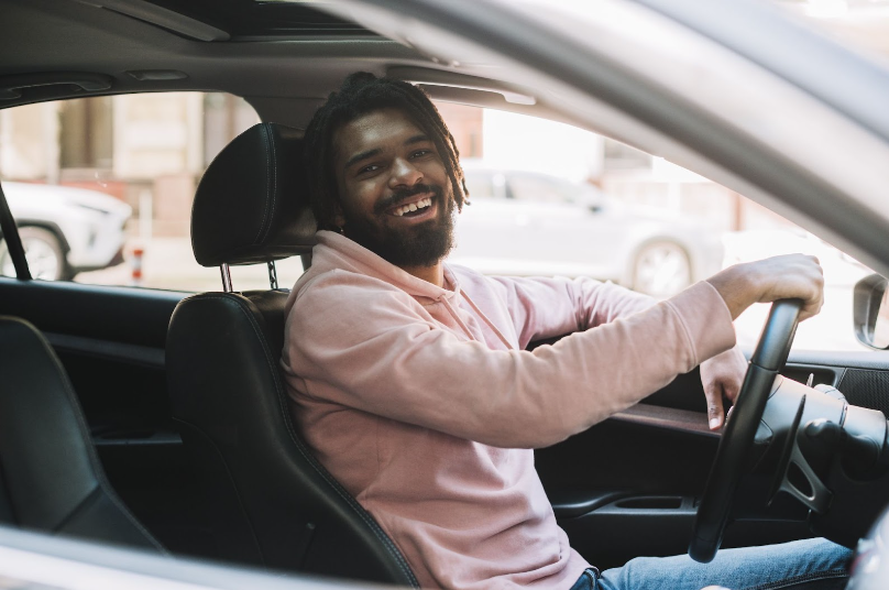Homem com dread e barba vestindo uma blusa rosa, sentado no banco do motorista com um braço apoiado na janela e a outra mão no volante, olhando para o lado e sorrindo.