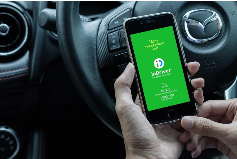 Mãos segurando um celular com o aplicativo InDrive aberto, com um volante ao fundo.
