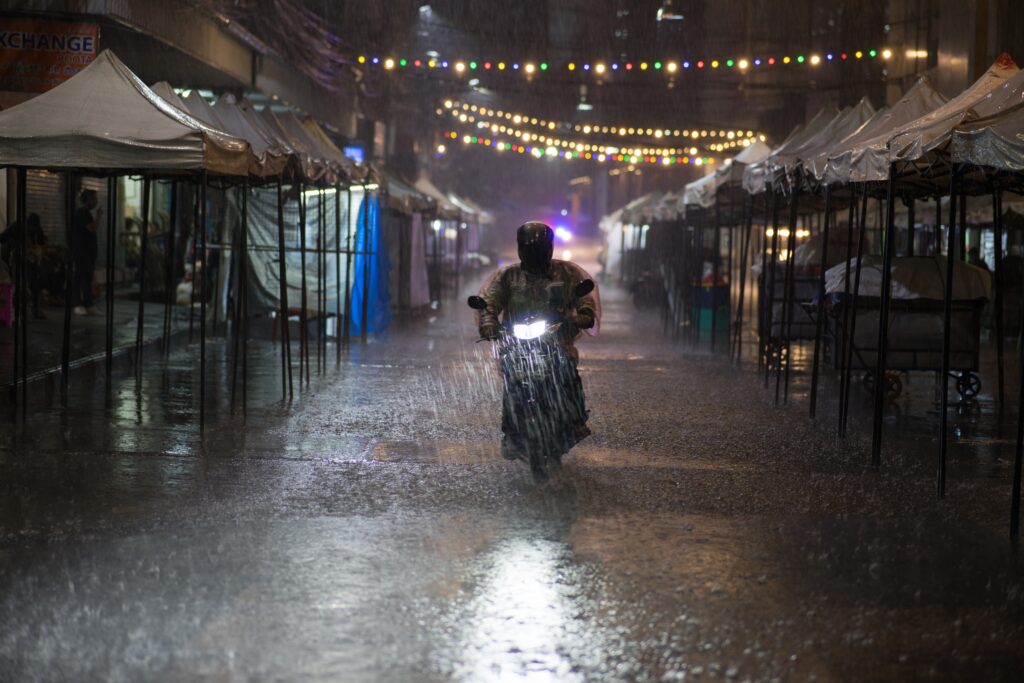 Motociclista dirigindo em meio a chuva forte, enquanto atravessa uma rua com luzes.