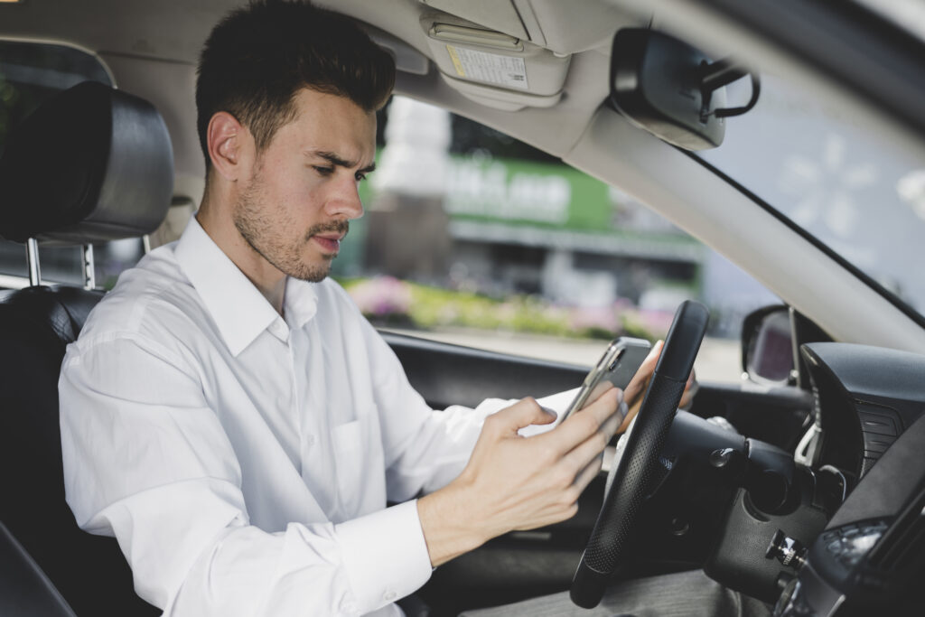 Homem de cabelo curto e camisa branca sentado no banco do motorista, com uma mão no volante e outra no celular, olhando para o celular.