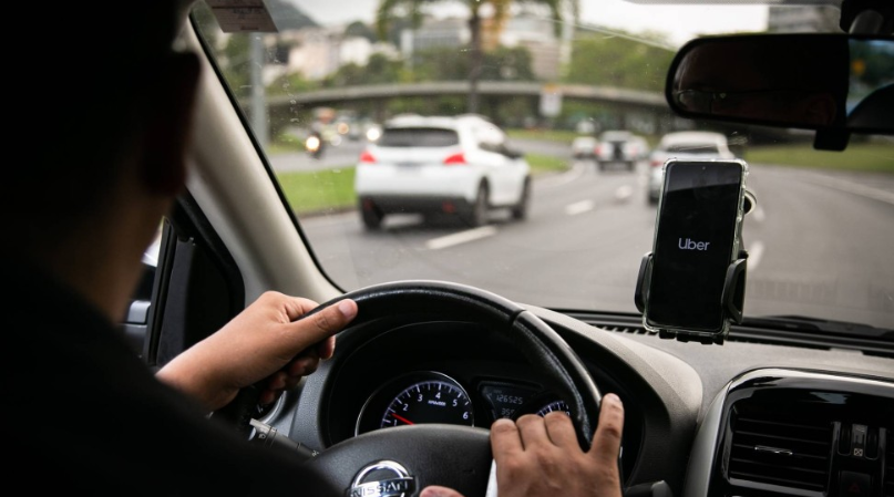 Homem de costas dirigindo um carro. No celular em um suporte no vidro do carro, a logo da uber.