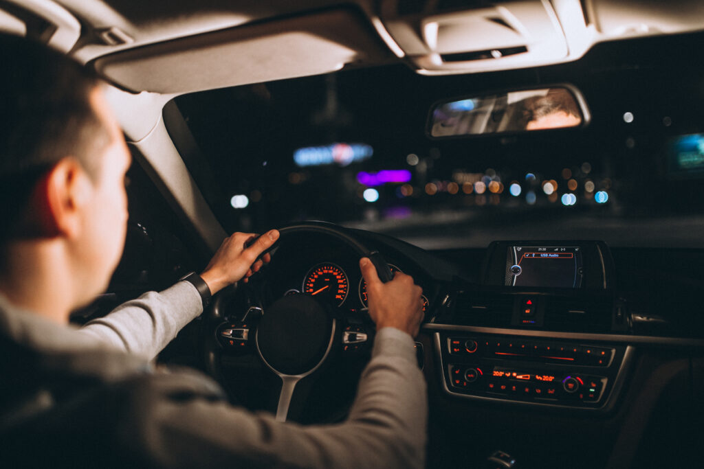 Homem de cabelo curto e blusa cinza de costas, sentado no banco do motorista com as mãos no volante, dirigindo um carro a noite.