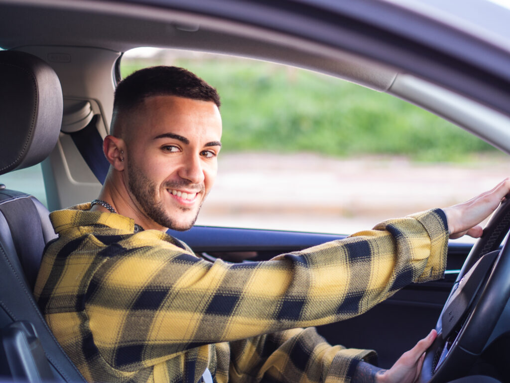 Homem de cabelo curto usando camisa xadrez amarela e preta sentado no banco do motorista com as duas mãos no volante e sorrindo.